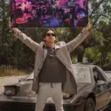 Man stands in front of car and holds large magenta and navy wall art overhead featuring abstract drawing of city street at night.