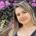 Female artist Karine Tonial Grimm stands outside in front of a deep purple flowering bush, with her long brown hair cascading around her. She smiles gently at the camera with a closed lip grin in this tranquil and natural setting.