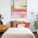 A light pink and yellow abstract artwork is hanging above a little girl's bed which is covered with a light pink duvet and several pink and yellow throw pillows. The artwork features shades of pink from light to dark mixed with mustard yellow and one accent of navy blue, adding a playful and vibrant touch to the room's décor.