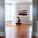 A girl sits on the floor in an empty blue living room, looking thoughtfully off into the distance while below a pink and blue abstract artwork with black accents, titled "Pink Horizon". The walls of the living room are painted light blue and the floor is a shiny walnut-colored wood, creating a serene and calming atmosphere in the room.