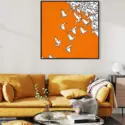 Boho modern art hung on a light grey wall above a large mustard colored couch. The boho art features a bright orange background with origami birds flying across