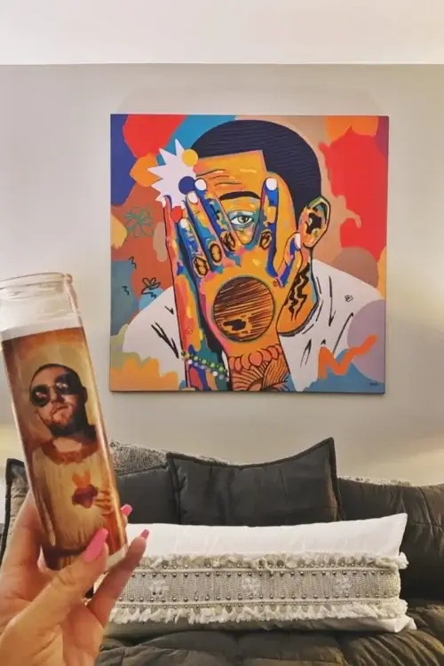 Massive colorful mac miller hip hop art in square hangs on white wall above grey couch in modern living room. A person holds a tall celebrity prayer candle of Mac Miller in front of the artwork, in tribute to the artist.