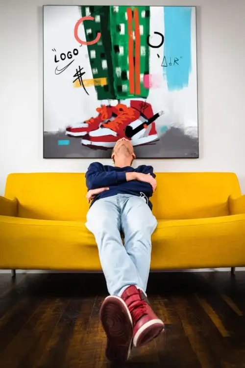Huge colorful hip hop art of digitally drawn Air Jordans on a person wearing green and white checkered pants hangs above yellow couch. On the couch, a man casually leans back, chin tilted towards the sky. On his feet are red Air Jordans, which are similar to the sneakers drawn in the wall art.