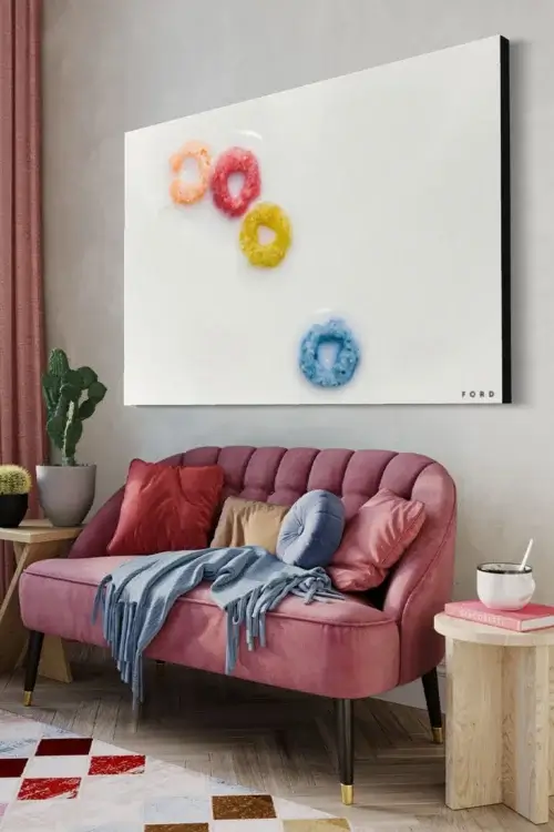 Huge minimalist wall art of 4 fruit loops - orange, red, yellow, and blue - floating in milk hangs on wall above pink couch decorated with blue throw pillows and blankets in boho living room.