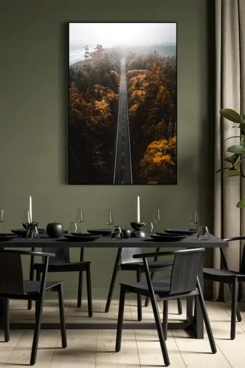 Large nature photo of road through forest in fall hangs on green wall in dinning room above black kitchen table.