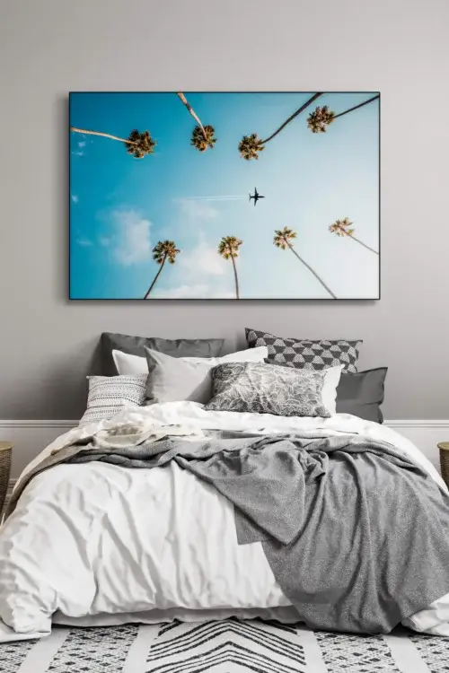 Photo featuring plane flying between palm trees through blue skies hangs above bed in bedroom decorated in neutral and grey.