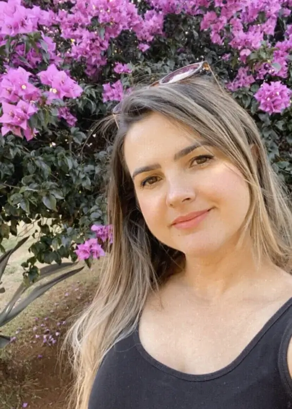Female artist Karine Tonial Grimm stands outside in front of a deep purple flowering bush, with her long brown hair cascading around her. She smiles gently at the camera with a closed lip grin in this tranquil and natural setting.