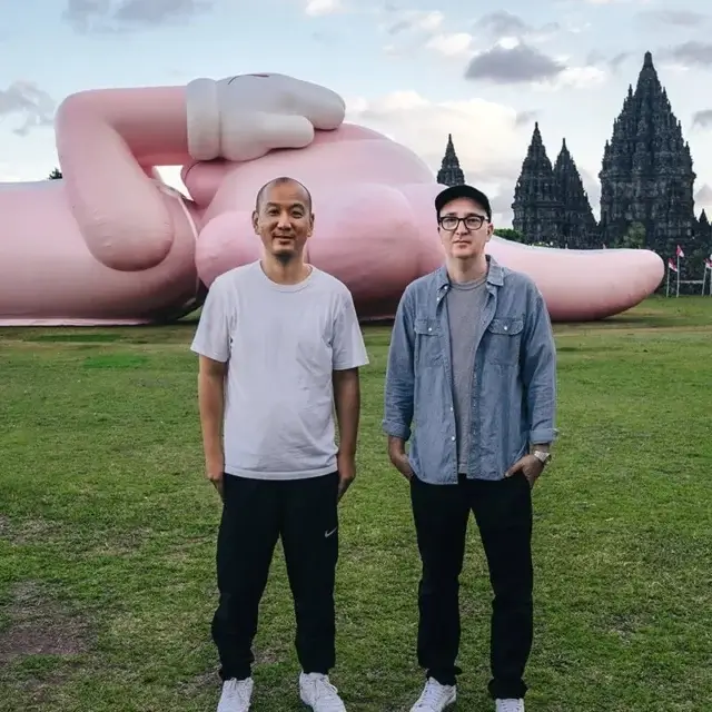 KAWS:HOLIDAY scuplture laying in the grassy lawn beside 9th century Indonesian temple, Prambanan Temple. KAWS and AllRightsReserved standing infront,