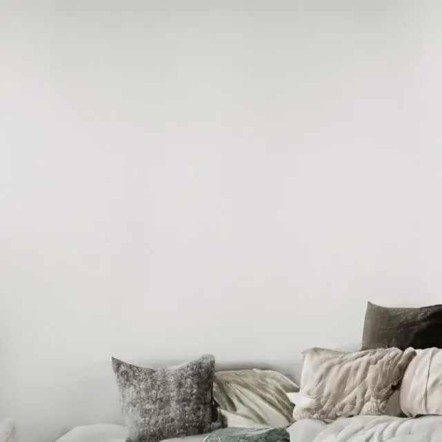 Neutral/white room with neutral and white furniture including a white couch with white and grey pillows. To the left is a green house plant and to the right is a bedside table with a small lamp and a bright red colored book. On the wall you can see a bright red piece of BIG Wall Decor artwork on the wall. The artwork is a bright and vivid red.