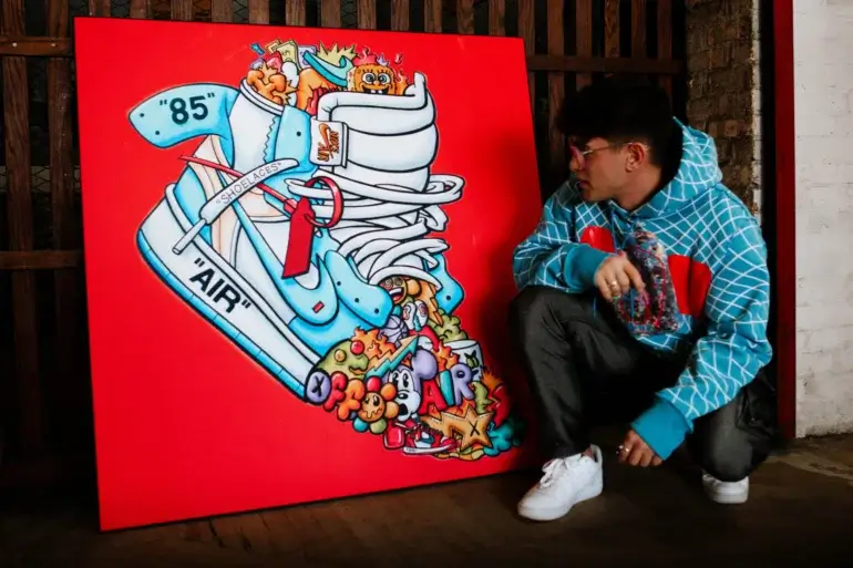 Man crouches next to large square pop wall art of blue Air Jordan sneakers with cartoon doddles coming out of toes and top of shoe.