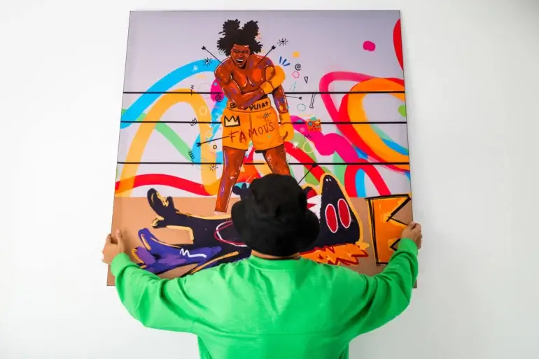 Man in lime green shirt hangs large colorful pop artwork of Basquiat fighting his monster onto white wall
