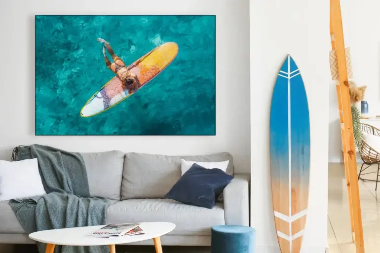 Aerial surfer girl photo taken in Hawaii as she drifts on her surf board in the ocean hangs on white wall above neutral couch in beachy living room.
