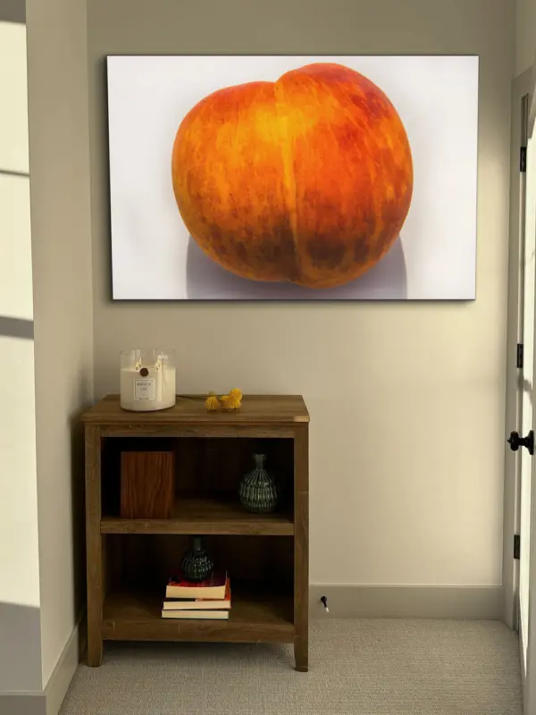 Large photo of a peach that mimics the shape of the peach emoji that is synonymous with a butt is shown on a white background and hangs on a neutral wall in a bedroom above shelf topped with Big Ass Candle in Sparkling Paloma scent.