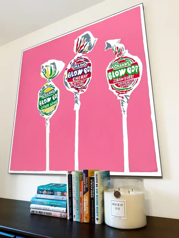 Large pink pop art featuring drawing of Blow Pops hang on bedroom wall above shelf with piles of books and a big ass candle