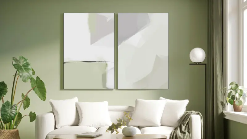 A modern, minimalist style living room with light sage green walls features two abstract pieces of art in light green, white, and light grey by artist Karine Tonial Grimm. The neutral color palette of the artwork complements the room's decor, which includes a white couch and a minimalist aesthetic.