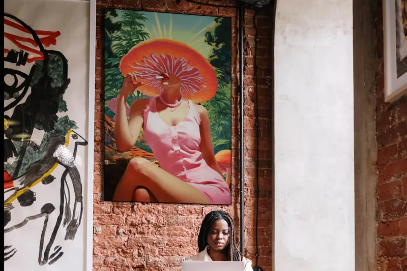 Large vintage mushroom art featuring woman smoking a joint hangs on brick wall above a person working at a desk in a stoner's apartment